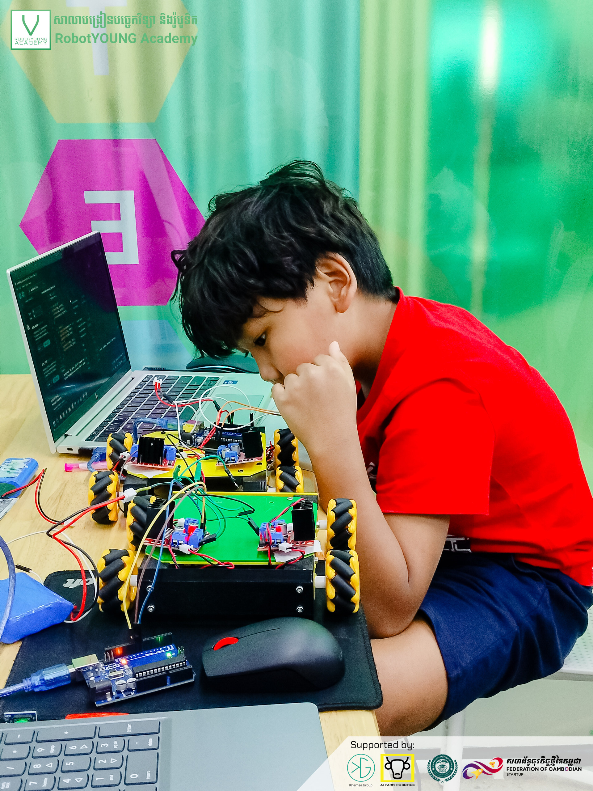 A student in a red shirt studying a wheeled robot beside an open laptop