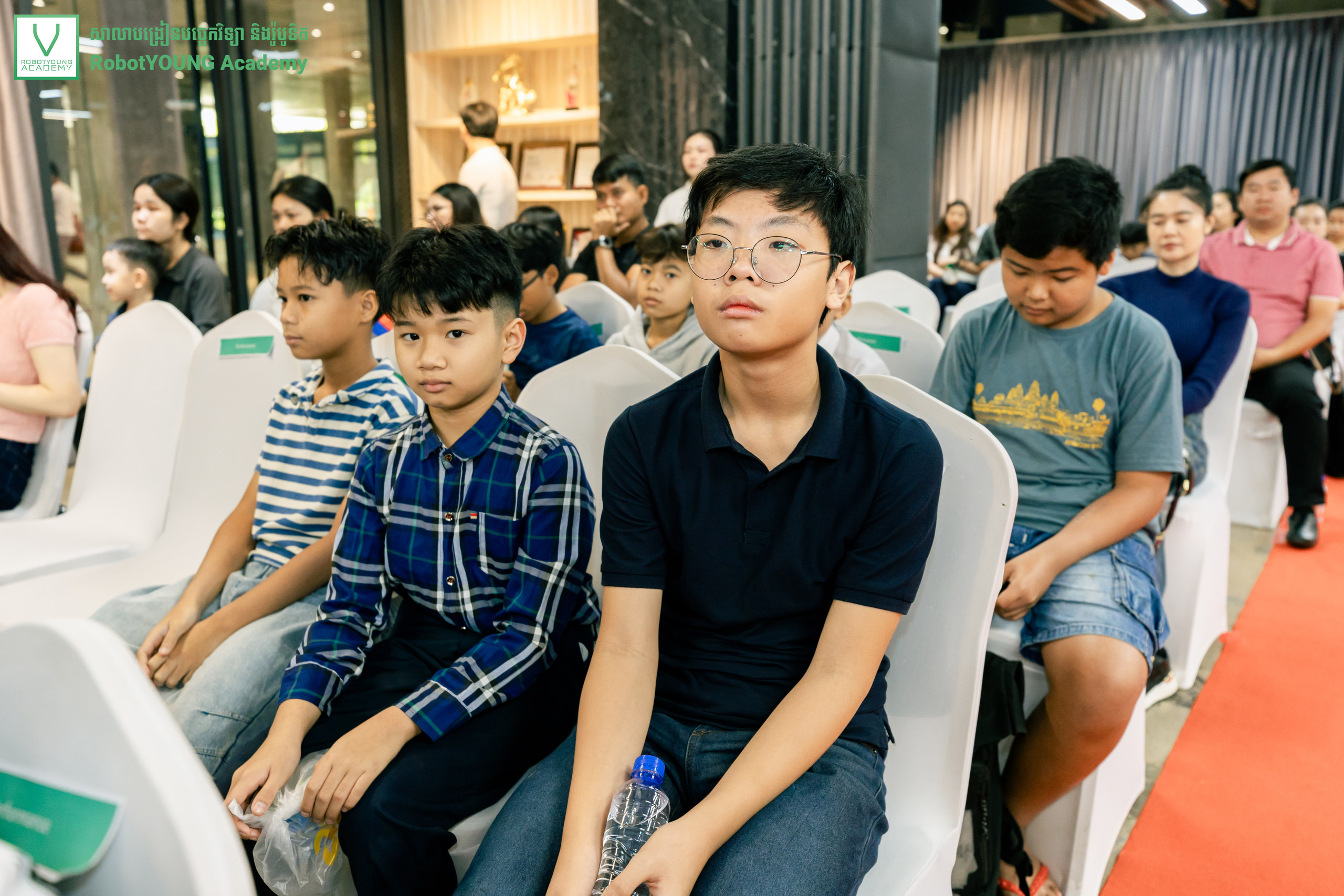 Children and adults seated in white-covered chairs at an academy event
