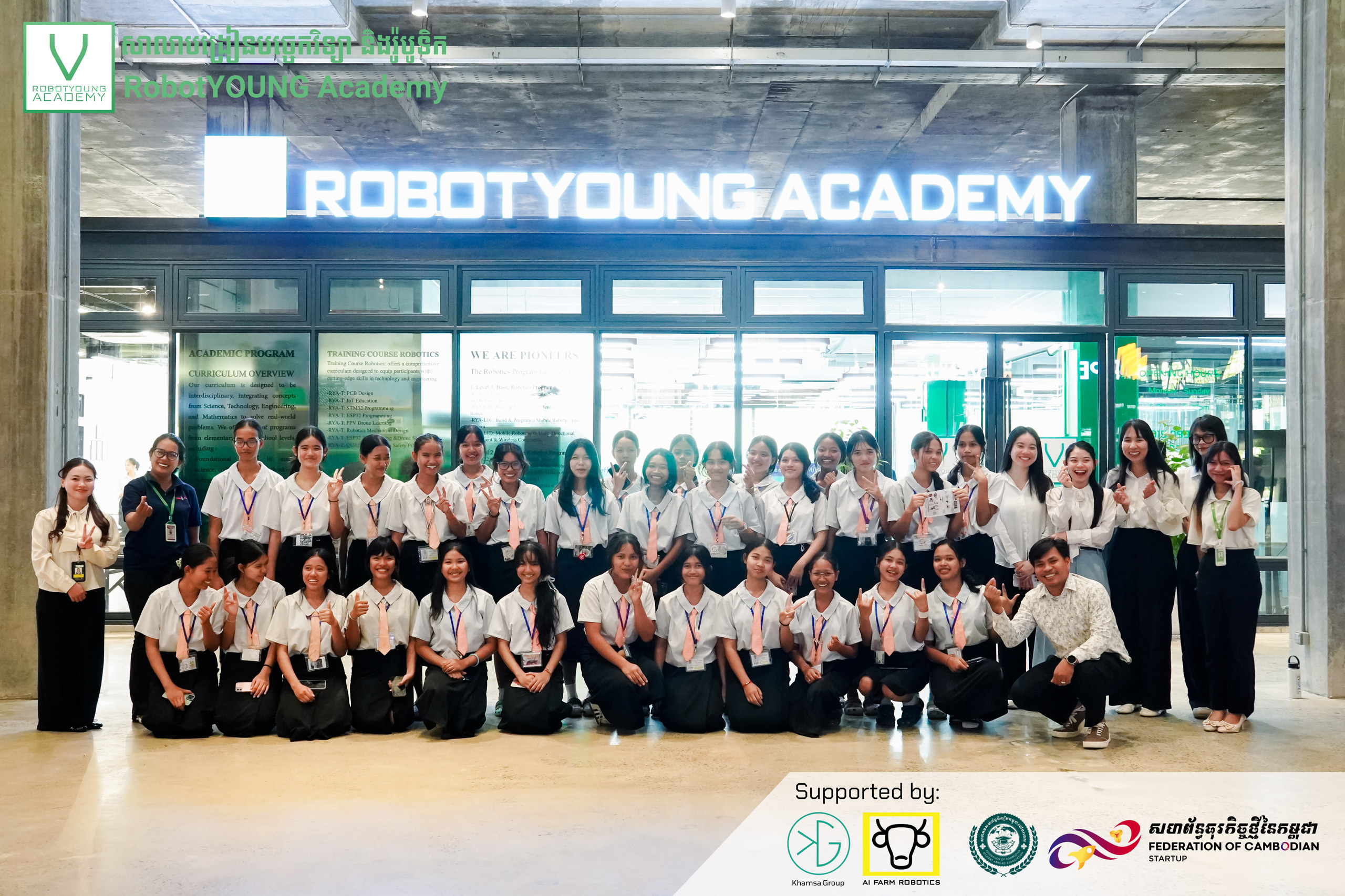 Students and adults posing together beneath the RobotYOUNG Academy entrance sign