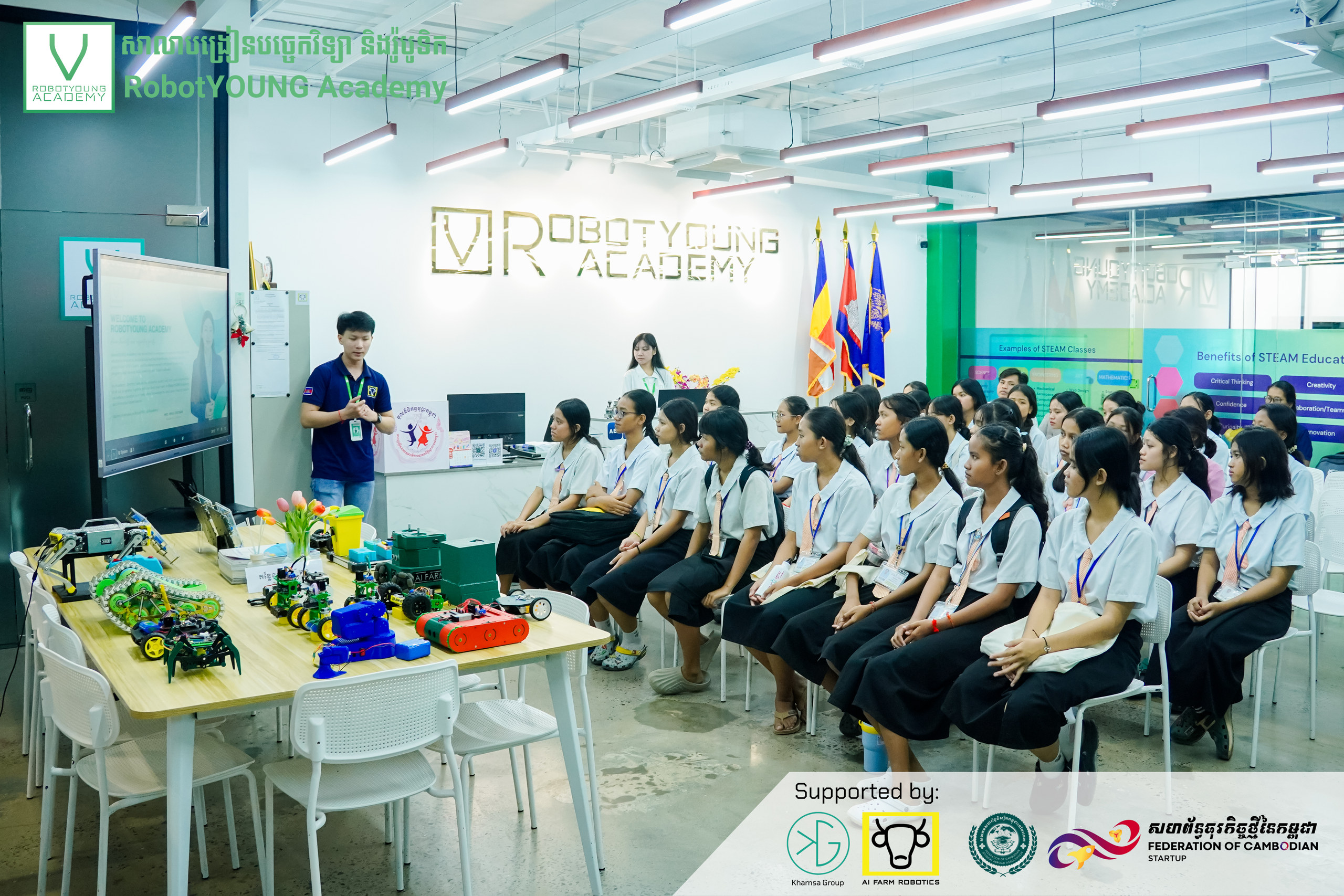 An academy guide presenting to seated students beside a table of colorful robots
